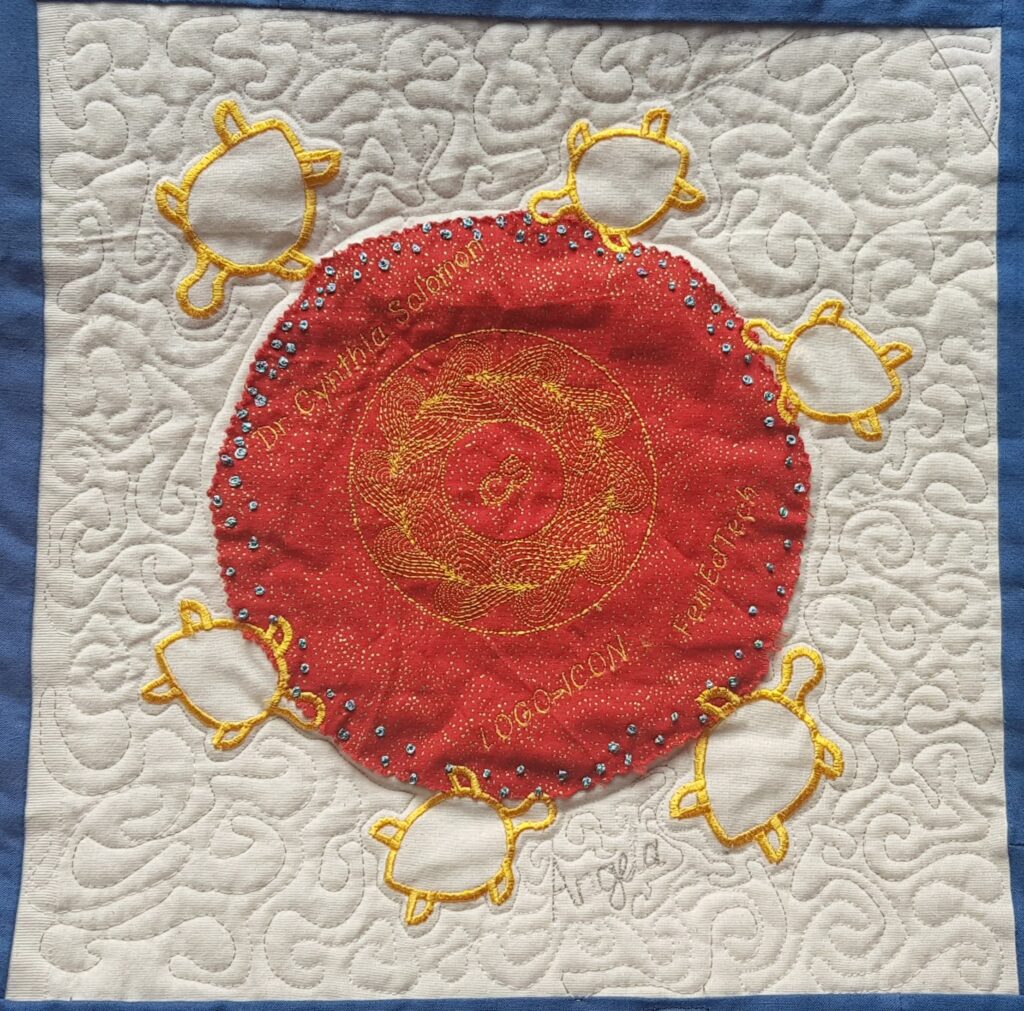 a square from a quilt, featured a central red circle, surrounded by yellow turtles, in a cream square. the red cirle had the text Cynthia Solomon, LOGO ICON, and the name Angela is stitched into the cream square.
