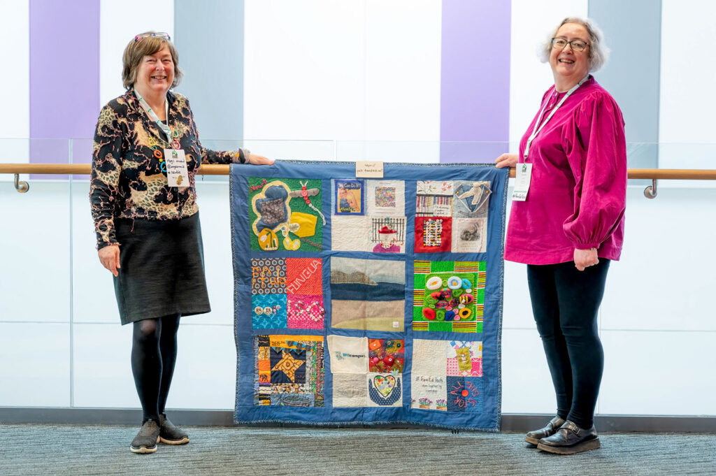 tow women stand smiling at the camera, holdin a quilt section about one square metre, multicolored, framed in blue