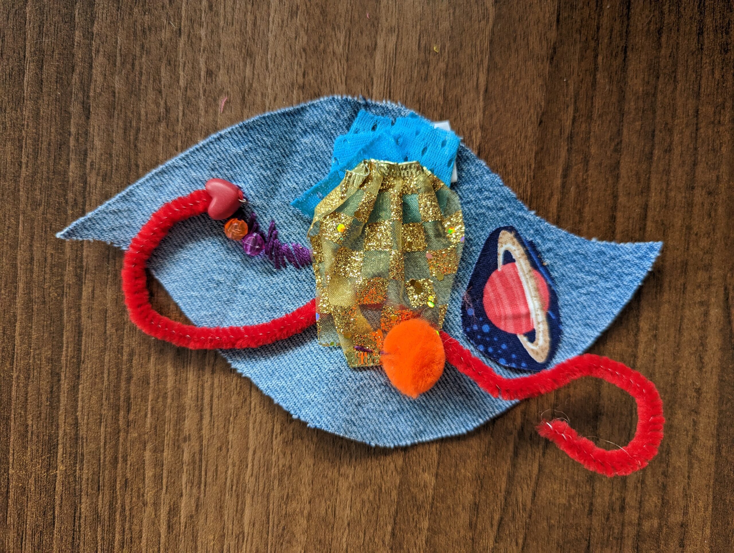 A leaf-shaped denim patch, with a purple piece of embroidery, red pipe cleaner, a patch with a planet with ring, a gold patch and orange pompom attached.