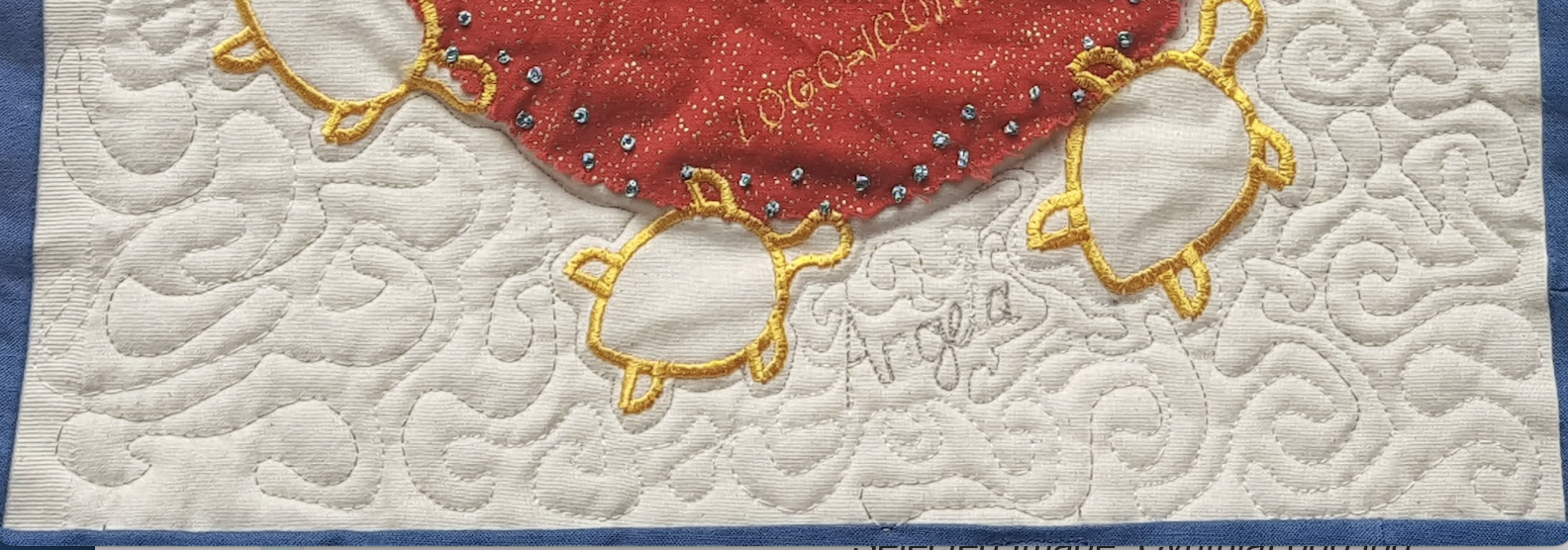 a close up on a square from a quilt, featured a central red circle, surrounded by yellow turtles, with the name ANGELA stitched into the cream square.
